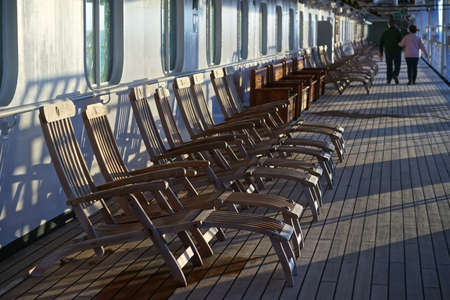 An elderly couple strolls past a long row of empty deck chairs on the Promenade Deck of a cruise ship.の写真素材