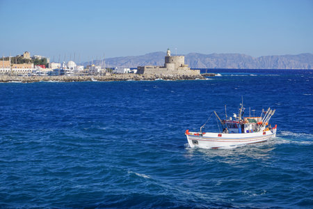 Rhodes, Greece: A fishing boat passes St. Nicholas Lighthouse (1863) in St. Nicholas Fortress in...