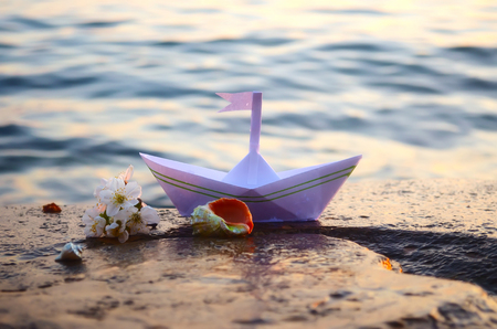 Paper boat, seashells and a sprig of cherry blossoms on a large stone on the seashoreの写真素材