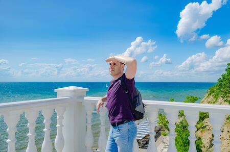 A healthy elderly man of athletic build stands near a white fence with balusters and looks at the blue sea.の写真素材