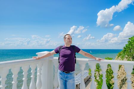 A healthy elderly man of athletic build stands near a white fence with balusters and looks at the blue sea.の写真素材