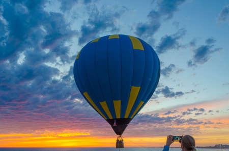 A girl takes pictures on her phone of a balloon at sunset in the sea. Air balloon in the evening sky.の写真素材