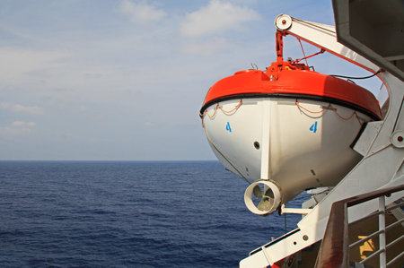 Red and white lifeboat on a passenger cruise ship hangs ready to launch next to a textured background of the calm open water of the Agean sea in Greece of the Mediterranean.の写真素材