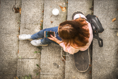 girl is sitting on a skateboard in headphones with a glass of coffee and a smartphone. View from aboveの写真素材