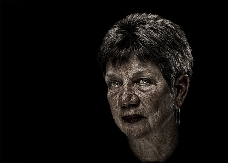 Portrait of a senior elderly older woman lady with short hair and desaturated with exaggerated flaws wisdom marks inlcuding wrinkles sunspots with serious judging melancholy sad depressed serious expression isolated on black backgroundの写真素材
