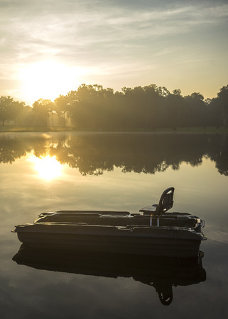 Small empty fishing boat on lake river water pond at sunrise sunset dawn early morning dusk with sun rays and trees forest on horizon feeling peaceful relaxed serene calm meditativeの写真素材