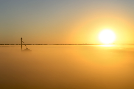 Agricultural farm building rising above fog on a foggy misty morning at sunrise or sunset in the rural countryside looking meditative relaxing calming beautifulの写真素材