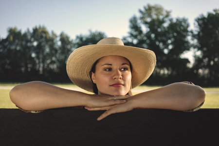 Cowgirl lady woman female wearing cowboy hat and flannel shirt with jeans leaning on country rural fence by a horse pasture paddock looking confident happy serene smart alone waiting watching patientの写真素材