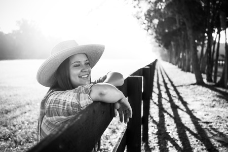 Cowgirl lady woman female wearing cowboy hat and flannel shirt with jeans leaning on country rural fence by a horse pasture paddock looking confident happy serene smart alone waiting watching patientの写真素材
