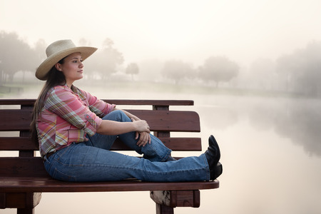 Girl woman lady female caucasian with long dark brown hair sitting on wood bench by foggy lake pond dock in cowboy hat flannel shirt looking relaxed happy serene beautiful young peacefulの写真素材