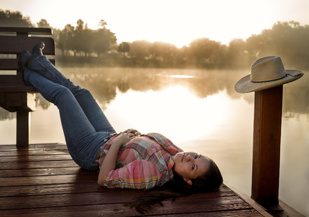 Girl woman lady female caucasian with long dark brown hair lying down on a lake pond dock with reflections at sunrise or sunset in cowboy hat flannel shirt looking relaxed happy serene beautiful young  peacefulの写真素材
