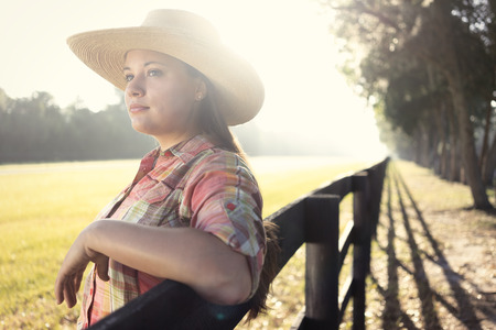 Cowgirl lady woman female wearing cowboy hat and flannel shirt with jeans leaning on country rural fence by a horse pasture paddock looking confident happy serene smart alone waiting watching patientの写真素材