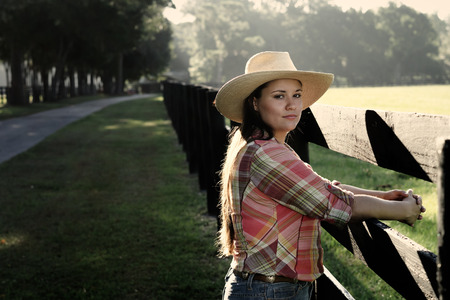 Cowgirl lady woman female wearing cowboy hat and flannel shirt with jeans leaning on country rural fence by a horse pasture paddock looking confident happy serene smart alone waiting watching patientの写真素材