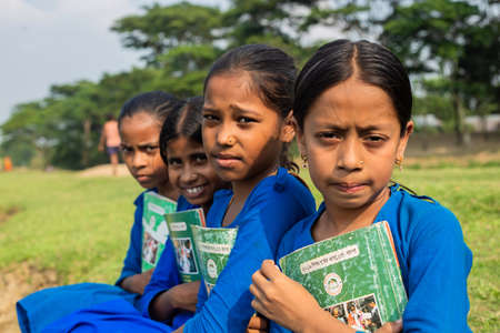 Gowainghat, Bangladesh â November 06, 2019: A group of school going girl students sitting in the school playground wearing school dresses and holding their books in the break time. Happy teen girls studying the booksのeditorial素材