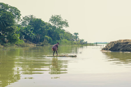 Taherpur, Bangladesh â November 05, 2019: Small boy drifting a raft by steering a paddle during flood in remote village. Little boy rowing handmade raft or boat made of banana trees tied together.のeditorial素材
