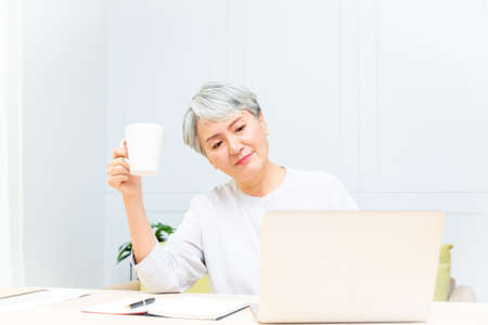 Senior asian woman at phone works on her laptop at home.