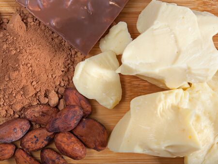 Pieces of natural cocoa butter, bar of milk chocolate, cocoa powder and cocoa beans on wooden cutting board. Top view. Flat layの写真素材
