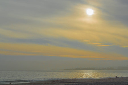 golden, smoky sky with sun shimmering on the ocean and a solitary person with a bicycle on a beach in Santa Barbara, Californiaの写真素材
