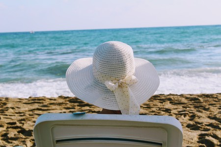 A woman sits in a deck chair against the background of a sea surf in a white hat with a wide brim.の写真素材