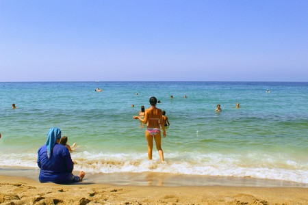 July, 2017 - A young girl in a bikini enters the sea. In the foreground, an elderly Muslim woman sits on the sand in the surf zone (Alanya, Turkey).のeditorial素材