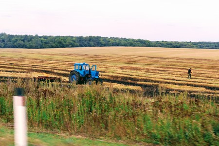 A blue tractor stands in the field against a background of forest and sky.の写真素材