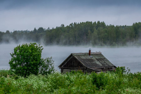 The traditional Russian log hut in poor condition on the bank of the river, early morning fogの写真素材