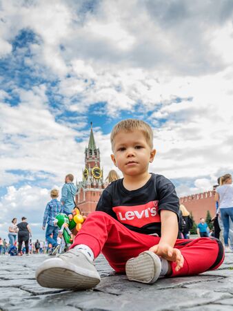 Moscow, Russia - July 7, 2018: little boy in red pants sits on Red Square paving stones, background from Spassky Tower, blue sky with clouds on sunny day. Tourist or political concept, oppositionのeditorial素材