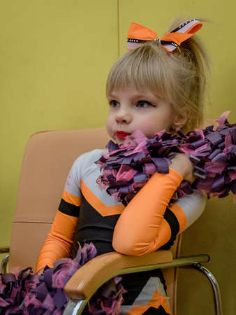 Moscow, Russia - December 22, 2019: Cute little girl cheerleader with pom poms, sits waiting for start of her performance, close-up portrait of a child athlete girl with blonde hair in sports uniformのeditorial素材