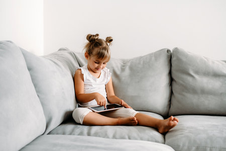 Toddler girl playing with digital wireless tablet computer on couch at home. Baby child growing with online applications. Child and electronic devices concept. Portrait of toddler with smartphone. Education and learning concept.の写真素材
