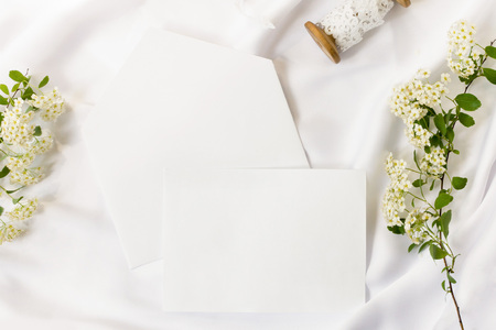 Flat lay workspace, mockup. Wedding invitation cards, craft envelopes,white flowers, green leaves and lace on white satin background. Top view.の写真素材
