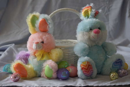 A couple of stuffed animal bunny rabbits with colorful eggs and a white basket between them.