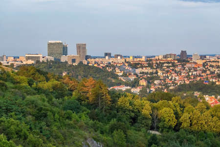 Prague, Czech Republic - August 21 2021: View of the city from distance with buildings and a skyscraper. Green trees in the foreground. Sunny summer evening, blue sky.の写真素材