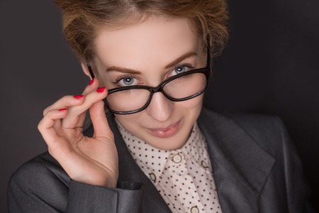 Red-haired business lady posing for photographer on dark background. Woman in grey business dress skewing at him so serious with her glasses dipped.の写真素材