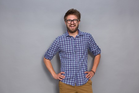 Portrait of smiling young man in glasses on grey background. Man in flowing navy blue plaid shirt posing with his arms akimbo.の写真素材