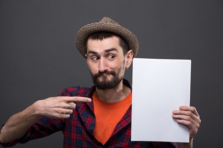 Portrait of serious hipster bearded man pointing out. Handsome man in hat keeping A4 sheet of blank paper in his hand.の写真素材