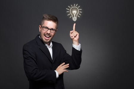 Happy businessman pointing out about tremendous idea on dark grey. Cute man in glasses showing to the bulb with his finger.の写真素材