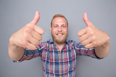 Smiling bearded man showing two thumb-ups. Happy man in navy blue, red plaid shirt satisfying his lifestyle on grey background.の写真素材