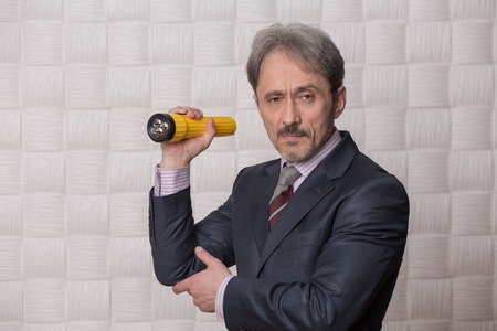 Mature businessman posing with yellow-coloured flashlight in his hand. Serious man is going to clear up the situation connected with low profit.の写真素材