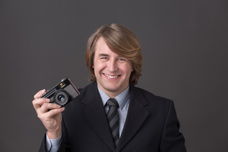 Smiling businessman keeping black camera isolated on dark grey. Man with blond hair in black business suit is very successful in company.の写真素材