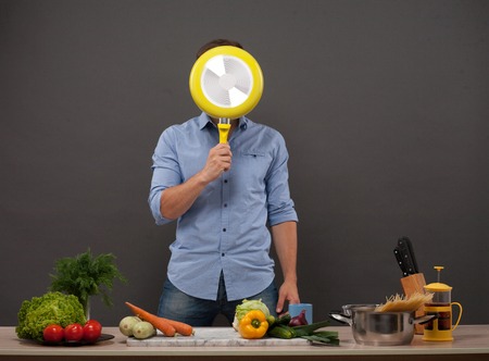Young chef hiding his face with yellow-coloured pan isolated on dark grey. Man in blue shirt posing in kitchen studio with many products on the counter.の写真素材