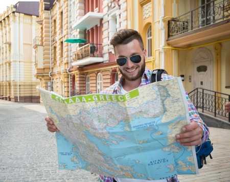 Smiling hipster tourist on the street looking at a guide. Bearded man in sunglasses with backpack...