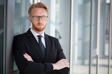 Portrait of serious businessman with folded arms posing near glass window. Blond handsome man in glasses looking away.の写真素材