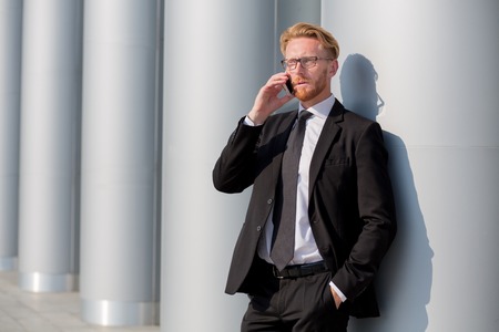 Elegant businessman in black suit speaking over mobile phone. Red-haired man in glasses discussing several isuues and problems.の写真素材