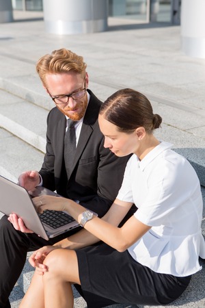 Business people sitting near office building with laptop. Smart businesswoman asking questions in his colleague near by.の写真素材