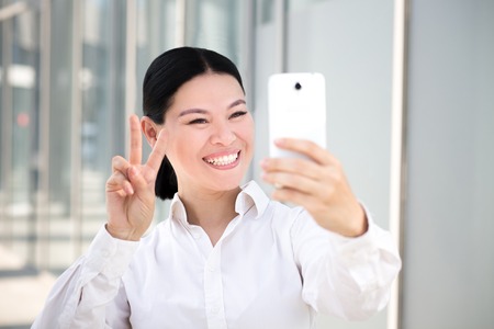 Young businesswoman making selfies on smart phone. Smiling lady in white shirt showing yo sign to the camera on her cell phone.の写真素材