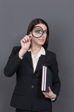 Portrait of business lady looking through loupe. Pretty woman in black business suit carrying documents and files in her hand isolated on grey.の写真素材