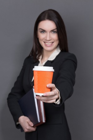 Business lady representing an orange cup of delicious coffee. Long-haired woman in black business suit smiling isolated on grey.の写真素材