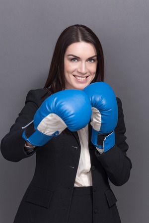Portrait of beautiful and young business woman wearing boxing gloves. Model in black business suit smiling and she is ready for battle.の写真素材