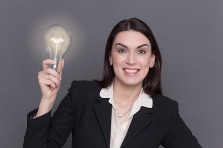 Business lady posing with bulb in her hand. Woman with long hair in black business suit happy smiling because of having new ideas.の写真素材