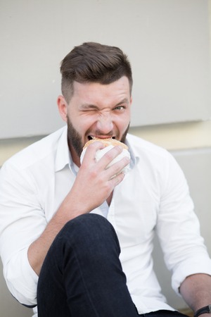 Handsome bearded man eating hamburger outdoors. He is hungry and having a good bite. Businessman resting during lunch time.の写真素材